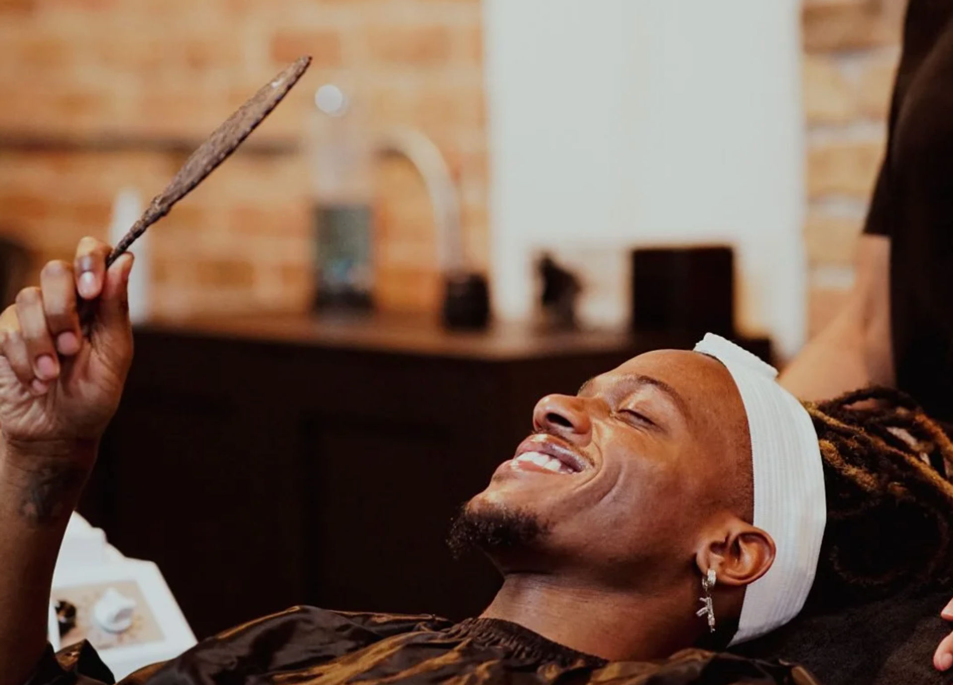 Smiling salon client reclining with a white headband, holding a handheld mirror to admire a fresh haircut in a warm, cozy grooming space.