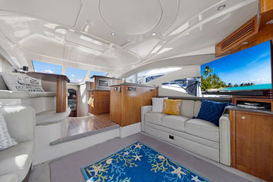 Sunlit luxury yacht salon with cream leather sofas, wood cabinetry, blue coral-pattern rug, colorful throw pillows, and a flat-screen TV showing a tropical beach through large windows with ocean views.