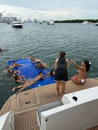 People relaxing on a large blue floating mat beside a boat swim platform, anchored yachts nearby and a coastal city skyline across the bay.