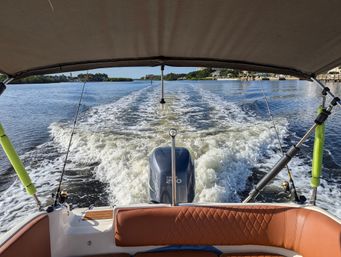 View from the stern of a motorboat under a bimini, showing an outboard engine, two fishing rods and foamy wake on a sunny coastal waterway with shoreline homes.