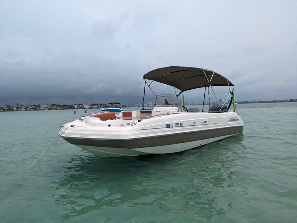 White motorboat with a dark bimini top anchored in clear turquoise bay near shoreline homes under an overcast sky, relaxed boat-day vibe