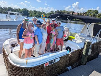 Smiling group of adults posing on a pontoon boat at a sunny Florida river dock, colorful beach ball, casual summer clothes and palm-lined shoreline in the background.