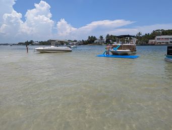 Shallow clear water at a sunny coastal sandbar with anchored leisure boats and a person climbing from a blue floating mat onto a pontoon, palm trees and waterfront homes under puffy summer clouds.