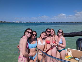 Group of six friends in colorful bikinis posing on a boat in clear turquoise bay on a sunny day, with a distant coastal city skyline and blue sky.