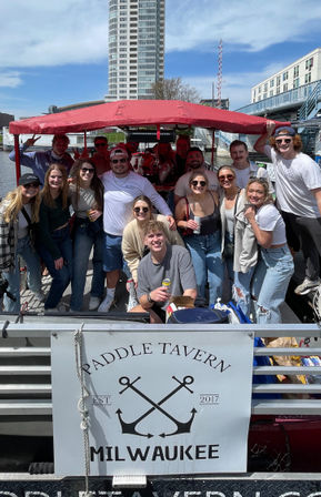 Group of young adults smiling and holding drinks on a red‑canopied party pontoon boat in Milwaukee harbor with city skyline and docks in the background.