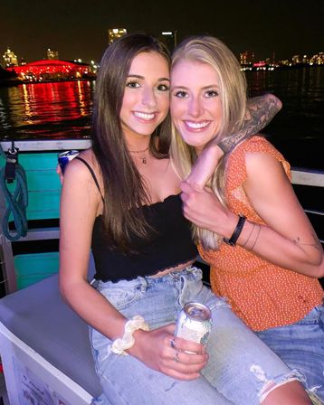 Two smiling friends embracing on a night boat ride, holding canned drinks, wearing casual summer outfits with a visible arm tattoo, waterfront city skyline and red-lit building reflecting on the water