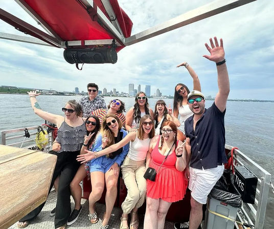 Smiling group of friends posing and waving on an open-deck boat tour with a distant city skyline and calm harbor water on a sunny day