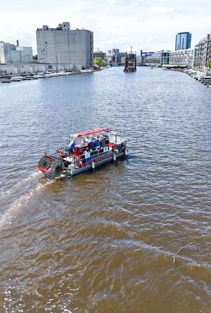 Red-and-gray paddlewheel boat with passengers cruising a wide urban river past marinas, industrial storage silos and waterfront apartment buildings under a bright sky.