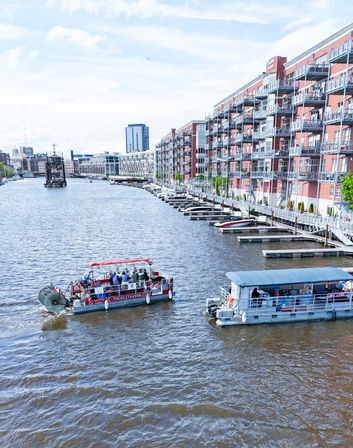 Urban riverfront scene with two tour boats cruising past red-brick waterfront apartments lined with balconies, private docks and a bright sky.
