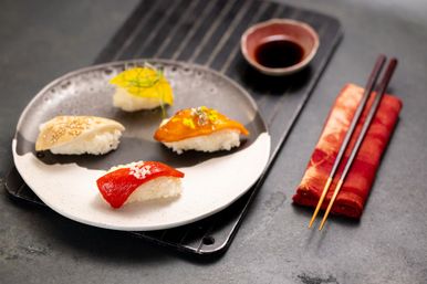 Colorful assorted nigiri sushi on a round ceramic plate with soy sauce dish and red chopsticks on a gray table — fresh Japanese restaurant-style presentation