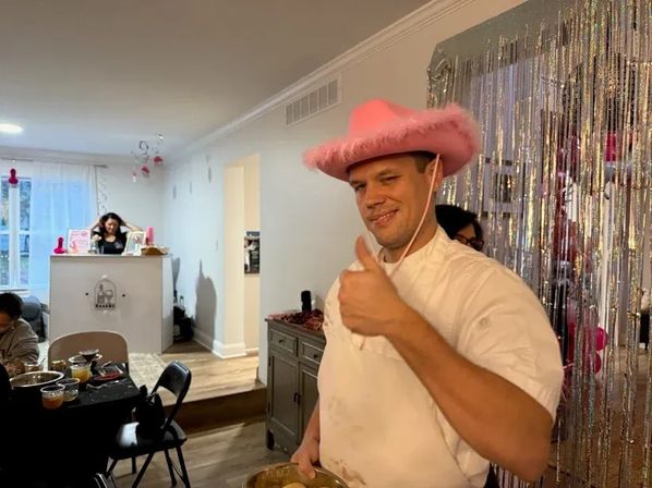 Man in a pink fuzzy cowboy hat giving a thumbs-up at a living-room party, holding a mixing bowl in front of a silver fringe backdrop with pink decorations and a snack table.