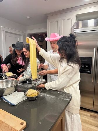 People in a home kitchen making homemade pasta — a long sheet of dough fed through a pasta machine on a flour-dusted island with bowls of dough and playful hats.