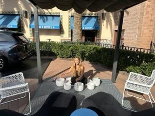 Person seated cross-legged under a shaded cabana in a sunlit outdoor mall courtyard, playing white crystal singing bowls on a rug, flanked by white metal chairs, hedges, parked car and blue storefront awnings.