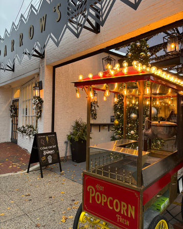 Vintage red popcorn cart with glowing bulbs outside a festive white-brick storefront adorned with Christmas wreaths and a lit tree.