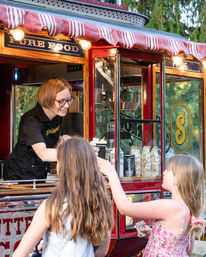 Smiling vendor hands popcorn to two kids at a vintage red popcorn cart with striped awning, glass displays and snacks at an outdoor summer fair