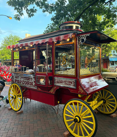 Bright vintage red-and-yellow popcorn cart with wooden wheels, striped awning and brass trim selling hot buttered popcorn on a tree-lined downtown brick sidewalk