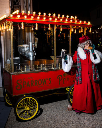 Festive nighttime street scene: person in a red coat with white-fur trim and plaid scarf and hat enjoys a snack while holding a popcorn box beside a red vintage popcorn cart lit by warm bulb lights.