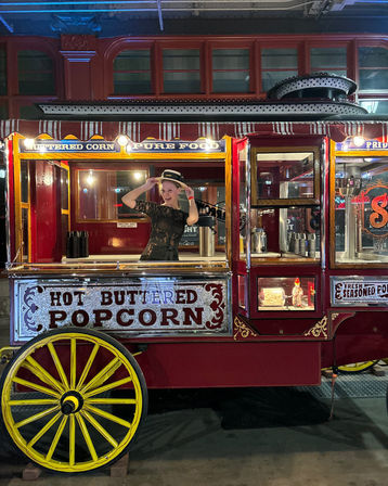 Person posing inside a red vintage popcorn cart with yellow wheel and illuminated “Hot Buttered Popcorn” sign at a nighttime street food / market scene.