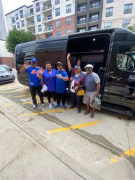 Five adults waving and smiling beside a black passenger van parked on a downtown sidewalk in front of modern apartment buildings, some wearing baseball jerseys.