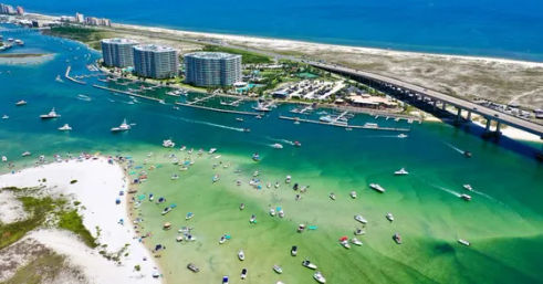 Aerial view of a sunny coastal inlet and barrier island with turquoise water, white-sand beach and sandbar dotted with anchored boats and swimmers, marina, curved bridge and high-rise condominiums along the shoreline.