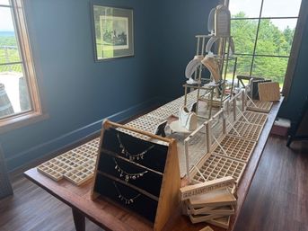 Sunlit wooden table jewelry display with necklace stands, ring trays and small purse mounts beside a large window overlooking green countryside