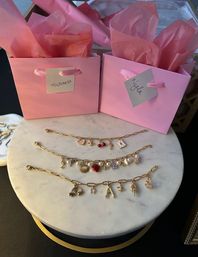 Three gold charm bracelets with pearls and enamel charms arranged on a round marble tray in front of two pink gift bags with tissue paper, ready for gifting.