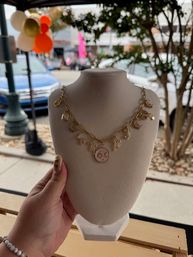 Gold charm necklace with moon-and-star enamel pendant on a beige jewelry display bust at an outdoor sidewalk market, parked cars and colorful balloons blurred in the background