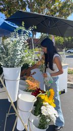 Shopper selecting sunflowers and mixed blooms (daisies, chrysanthemums, eucalyptus) from white buckets at an outdoor farmers market flower stand under a black umbrella