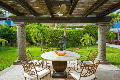 Shaded tropical garden patio beneath a timber-and-stone pergola with a round marble dining table, orange decorative bowl, six wrought-iron cushioned chairs, central stone fountain and palm trees on a manicured lawn.