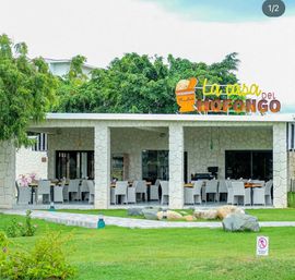 Outdoor dining patio at a tropical stone pavilion — rows of white wicker chairs and wooden tables on a manicured green lawn, leafy trees behind and a colorful restaurant sign above.