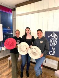 Three women in a bright craft studio holding decorated cowboy hats — two white hats with floral and western designs and one bright pink hat, standing on a wooden floor against a white shiplap wall with a navy seahorse painting in the background.