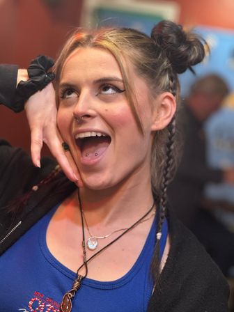 Playful close-up portrait of a young woman with space-buns and braids, septum and double-tongue piercings, bold winged eyeliner, wearing a blue tank top and layered necklaces, smiling animatedly in a lively indoor setting