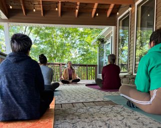 Outdoor yoga and meditation class on a covered wooden deck overlooking green trees, participants seated on mats under string lights and a hanging plant.