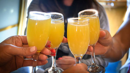 Four hands clinking champagne flutes filled with bright orange mimosas in a festive brunch toast