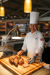 Smiling chef in white uniform and tall hat carving roast beef on a wooden cutting board at a restaurant carving station under warm pendant lights