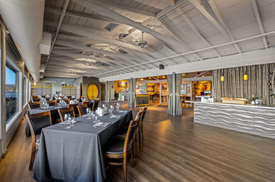 Spacious waterfront restaurant interior with long tables draped in black linens, wooden chairs, large windows overlooking a harbor, white exposed-beam ceiling, and a modern wavy-front bar with pendant lights.