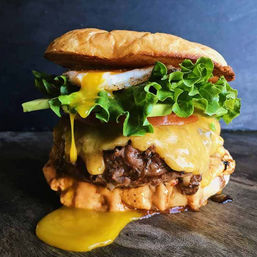 Close-up of a mouthwatering gourmet cheeseburger with melted cheddar over a juicy beef patty, crisp green lettuce and tomato, runny fried egg yolk dripping onto a toasted bun and creamy sauce on a wooden board