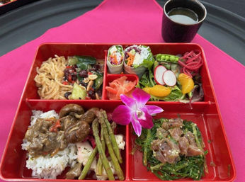 Red bento box lunch with Asian-fusion dishes: white rice topped with mushrooms and savory sauce, grilled asparagus, tuna poke on seaweed salad, cold sesame noodles, fresh vegetable spring rolls, mixed greens with radish and mandarin slices, pickled ginger, purple orchid garnish and soy dipping cup on a pink placemat.