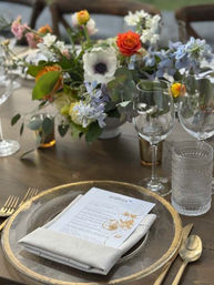 Elegant rustic wedding tablescape on a wooden reception table with a gold-rimmed charger, folded linen napkin and printed menu, gold flatware, textured water glass and wine glasses, and a colorful floral centerpiece with roses and anemone.