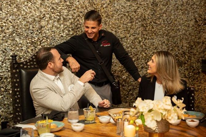 Two diners at a wooden table smile as a friendly server leans in to chat in an upscale restaurant with a shimmering gold mosaic wall; table set with cocktails, small plates, a lit candle and orchid centerpiece.