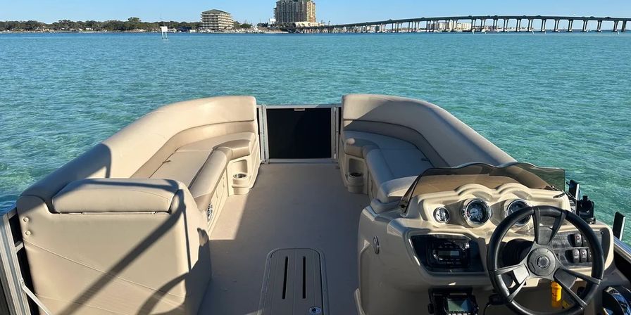 Sunlit pontoon boat interior with beige helm, steering wheel and gauges, wraparound bench seating and open deck on clear turquoise bay, coastal skyline and long bridge in the distance