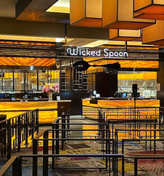 Glowing amber-lit hotel buffet counter with backlit stone panels, metal queue barriers in the foreground, geometric hanging lights, and a vase of pink flowers on the service counter.