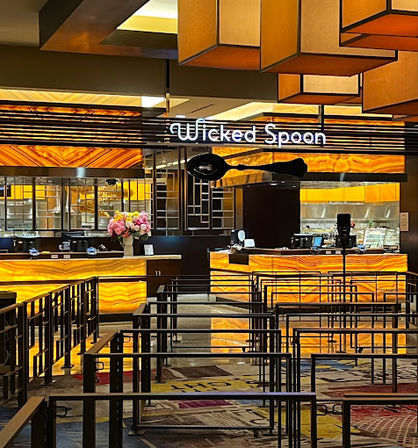 Glowing amber-lit hotel buffet counter with backlit stone panels, metal queue barriers in the foreground, geometric hanging lights, and a vase of pink flowers on the service counter.