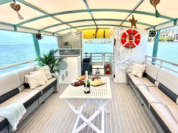 Sunny pontoon boat lounge on a blue bay with a coastal skyline and bridge in the distance — cushioned wraparound sofas, decorative pillows, potted palm, life ring, and central table with champagne and appetizers.
