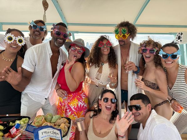 Group of friends at a sunny boat party by an urban skyline, wearing playful novelty sunglasses and summer outfits, laughing, holding drinks and enjoying colorful snacks.