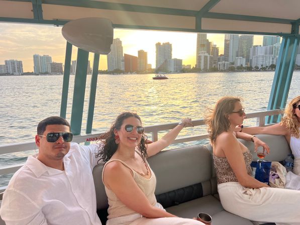 Group of friends lounging on a sunset boat cruise with Miami skyline and golden light reflecting on the bay.