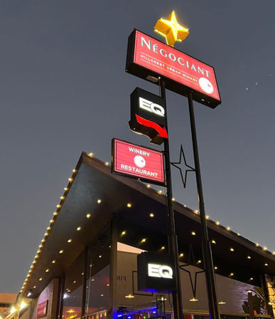Dusk city scene: illuminated urban winery and restaurant exterior with tall neon signs, glowing star topper and string lights along a modern canopy.