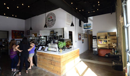 Sunlit coffee shop interior with rustic wood service counter, pastry display, syrup bottles, hanging pendant lights and customers ordering at the counter