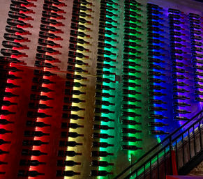 Wall of horizontally mounted wine bottles in neat rows, backlit by red, yellow, green and blue LED lights forming a rainbow gradient in a dim bar or restaurant interior.