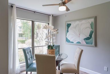 Bright dining nook with round glass table, tufted teal and cream chairs, woven-pot white orchid centerpiece, large window overlooking palm trees, floral wall art and ceiling fan.